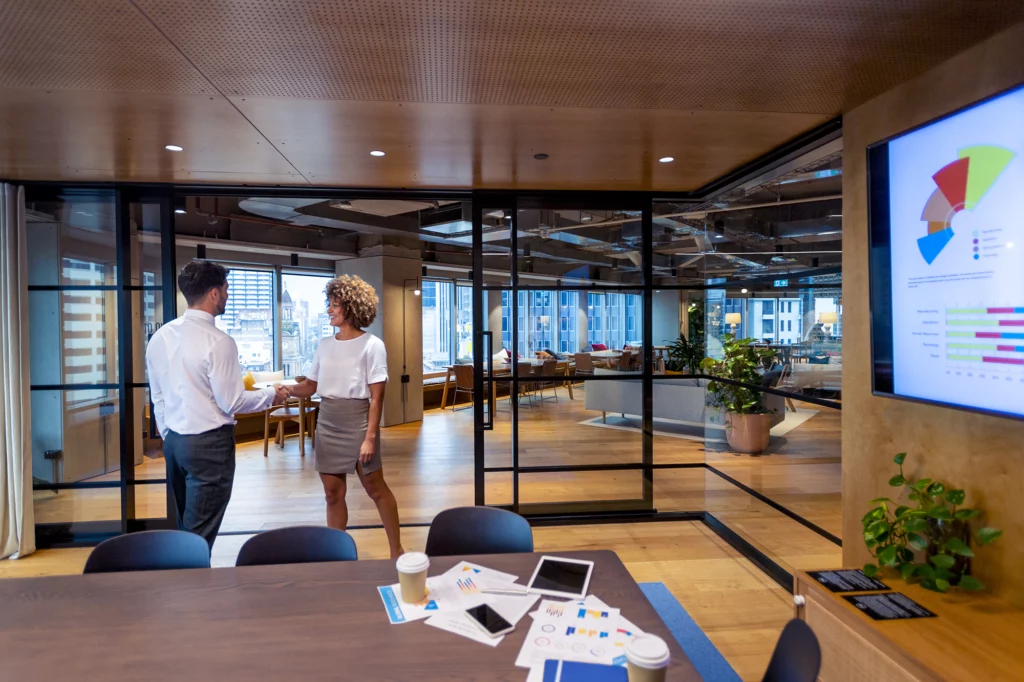 Man and woman handshake in a board room. Offices can be seen in the background.