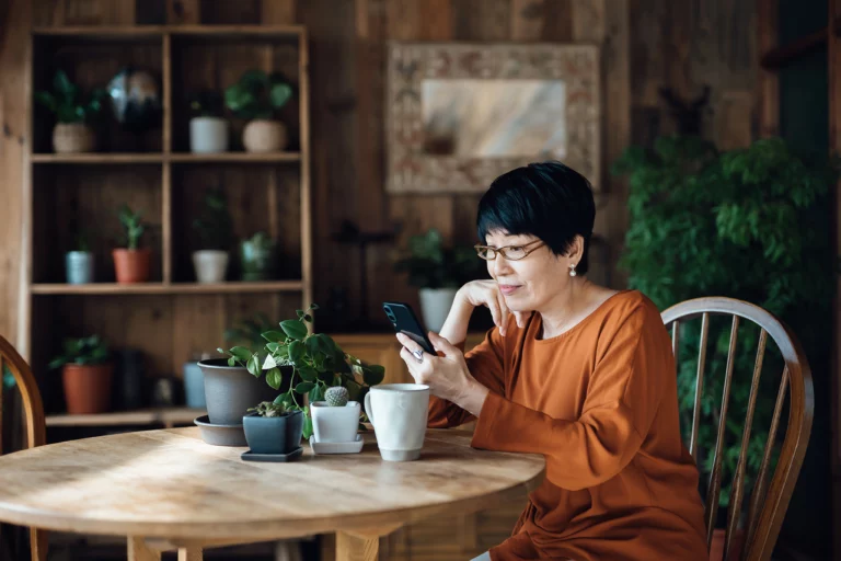 Smiling senior Asian woman sitting at the table, surfing on the net and shopping online on smartphone at home.